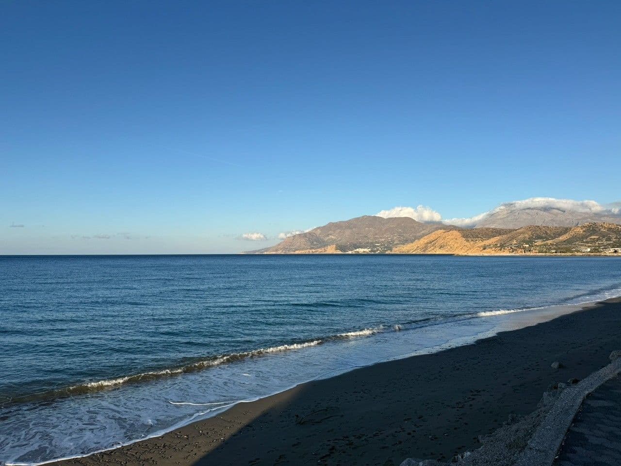 A long dark-sand beach, calm blue sea and the hills behind Agia Galini under a clear sky